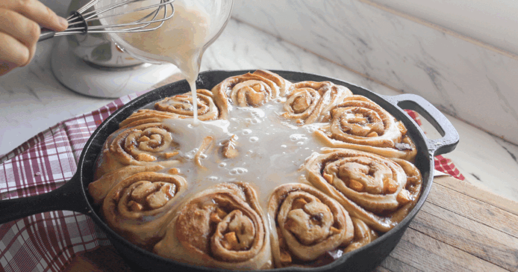 A stream of icing being poured onto freshly baked peach cinnamon rolls.