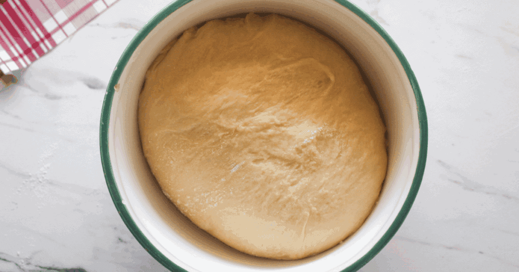 Soft peach cinnamon roll dough rising in a bowl during the proofing stage.