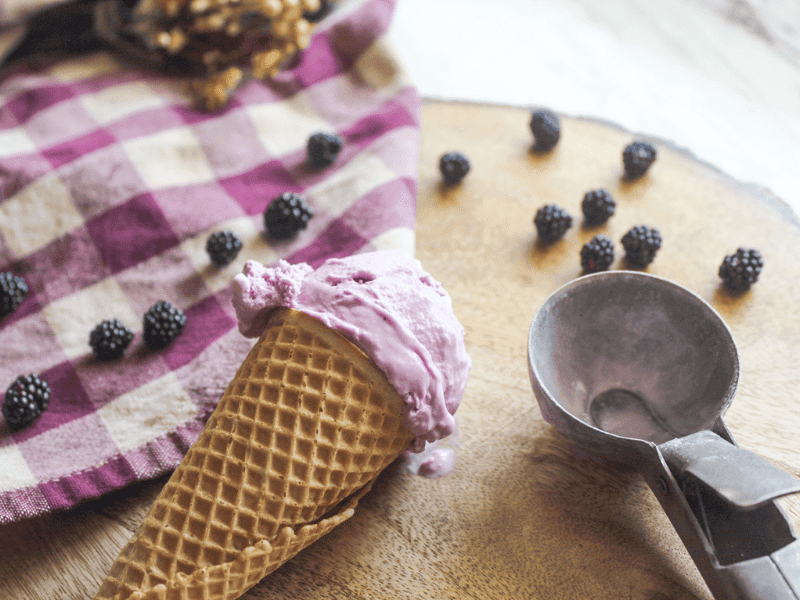 Blackberry ice cream cone resting on rustic wooden surface with berries scattered