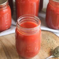 Open jar of roasted tomato sauce on a wooden board with sealed jars behind it, made using water bath canning