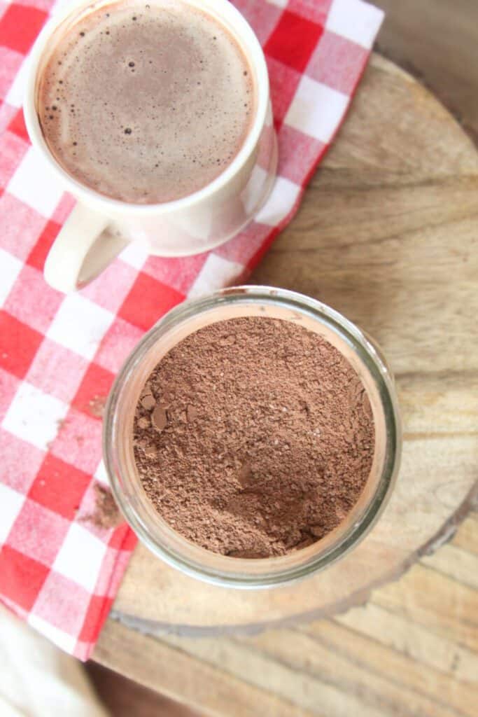 Homemade Hot Chocolate Mix (No Powdered Sugar!) Jar of homemade hot chocolate mix beside a mug of steaming cocoa on a red gingham napkin.
