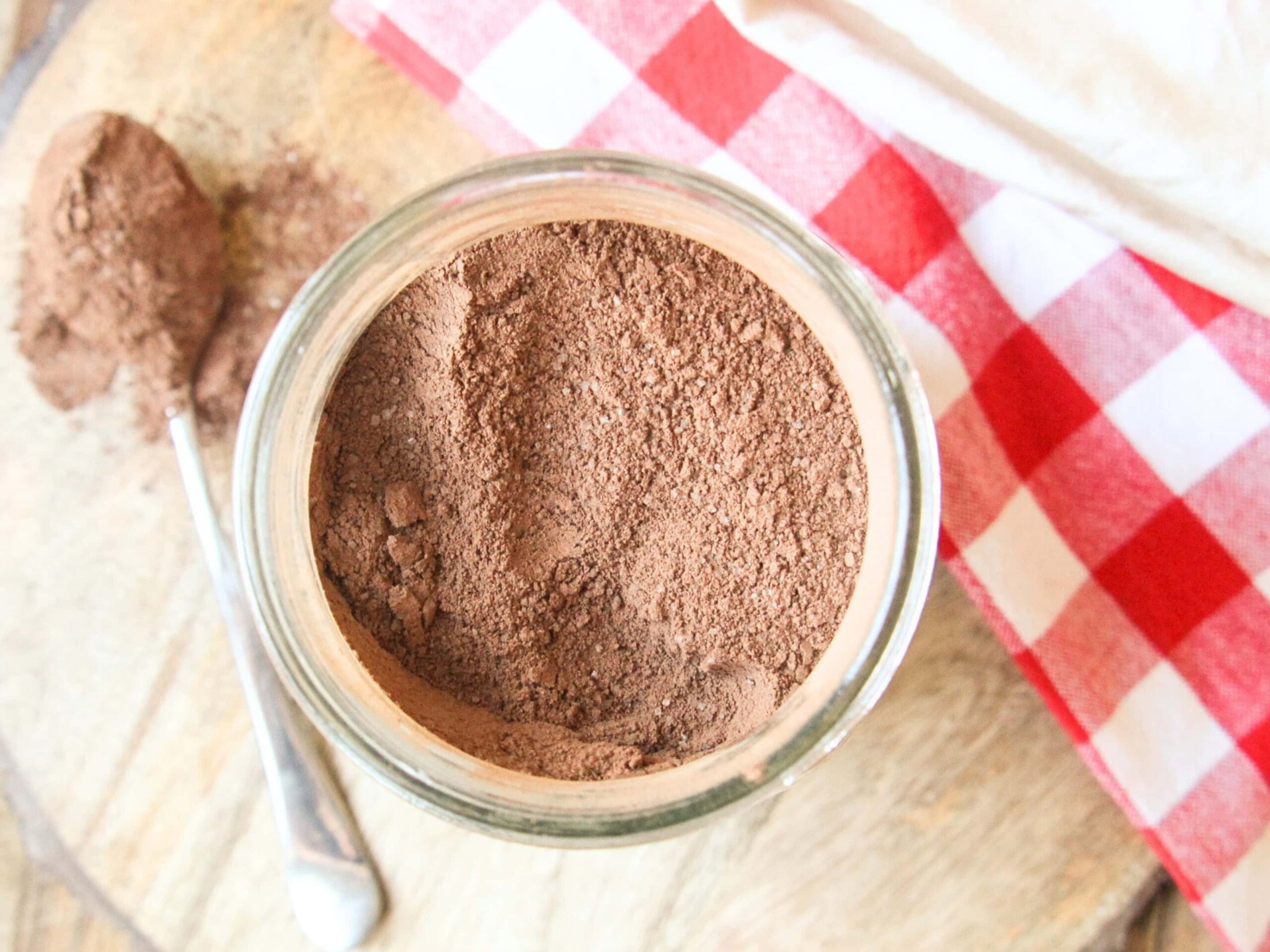 Homemade Hot Chocolate Mix (No Powdered Sugar!) Glass jar of homemade hot chocolate mix on a wooden board with cocoa powder spilled and a spoon.