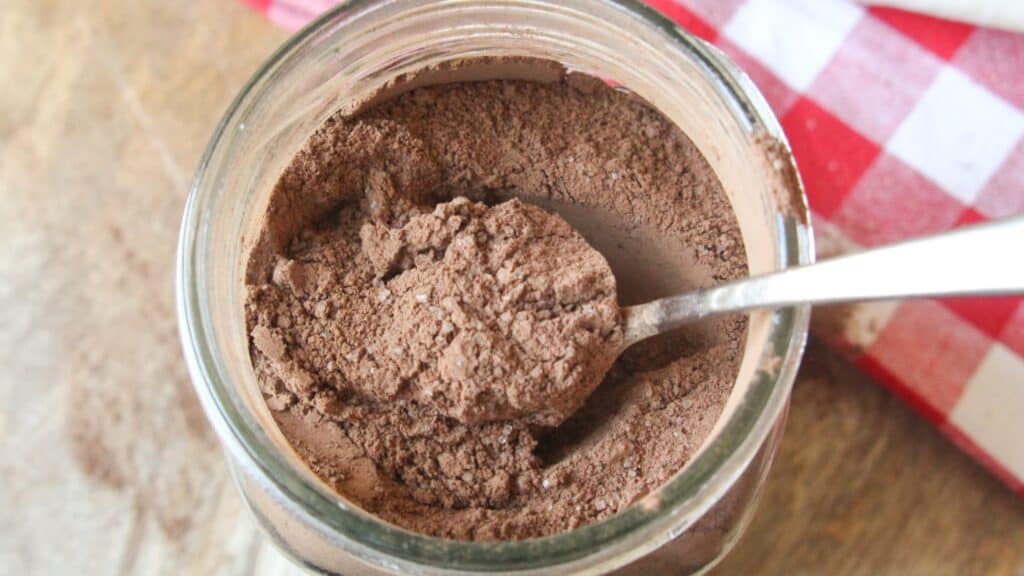 Homemade Hot Chocolate Mix (No Powdered Sugar!) Glass jar filled with homemade hot chocolate mix and a spoon scooping cocoa powder on a rustic wood board.