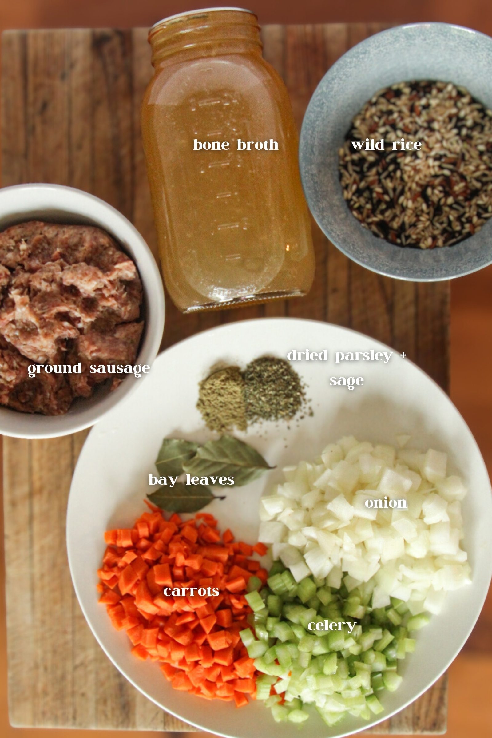 Flat lay of ingredients for sausage wild rice soup including ground sausage, wild rice, bone broth, carrots, celery, onion, parsley, sage, and bay leaves.