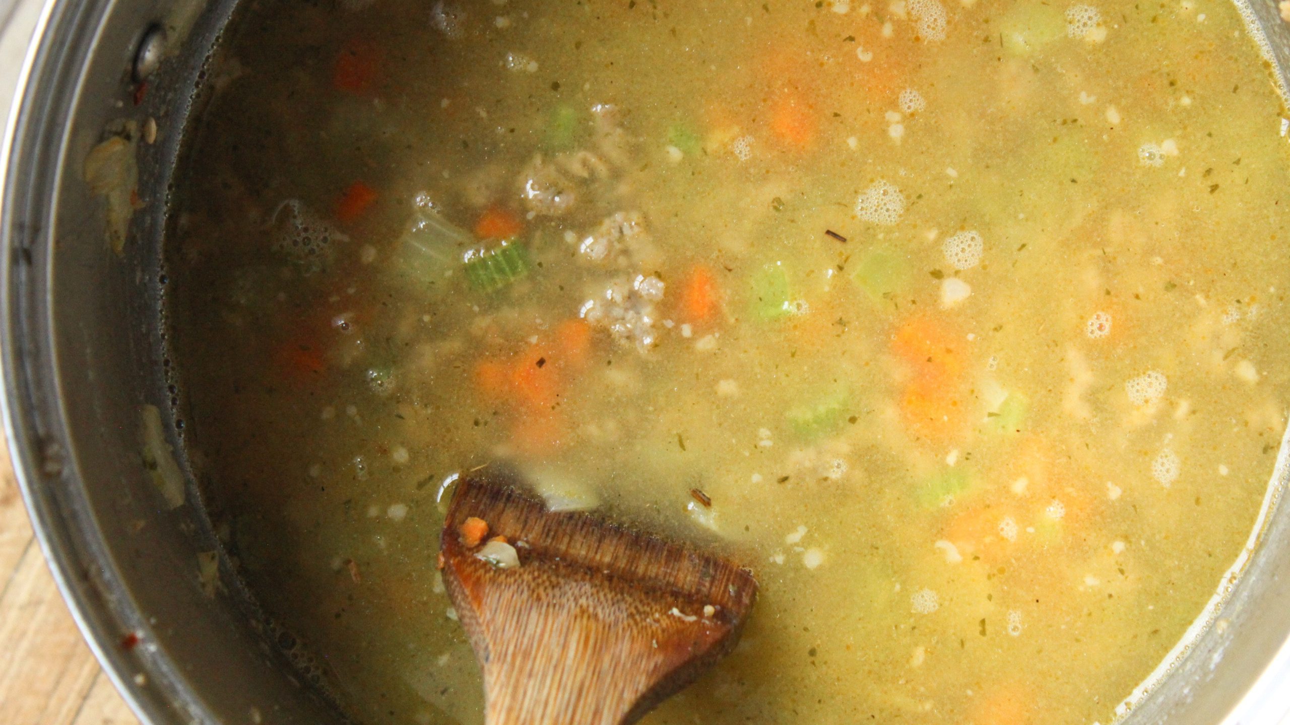 Sausage wild rice soup simmering in a pot with carrots, celery, and broth, stirred with a wooden spoon.