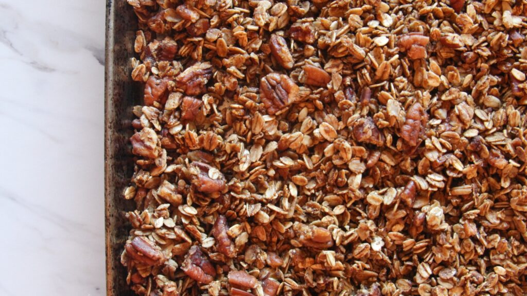Homemade pecan pie granola with maple syrup and toasted pecans on a baking sheet.