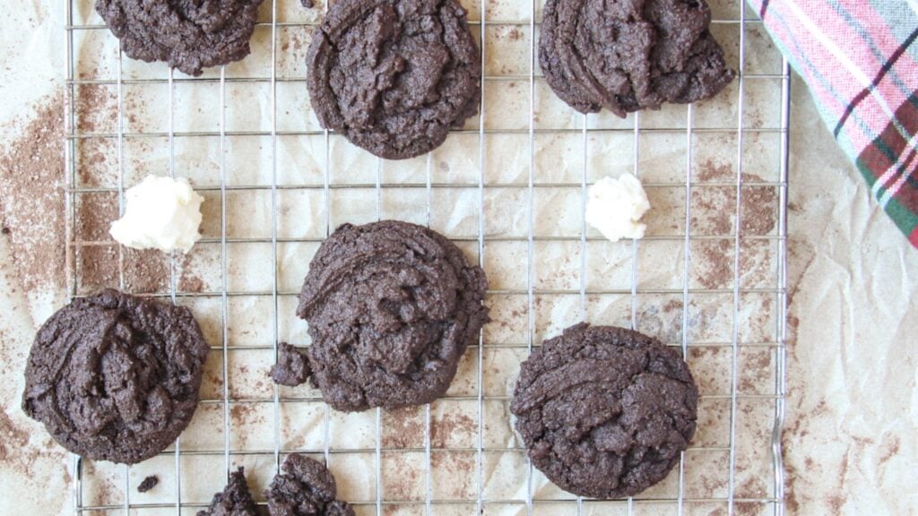 A batch of peppermint hot cocoa cookies with marshmallow pieces on a cooling rack beside a holiday plaid towel.