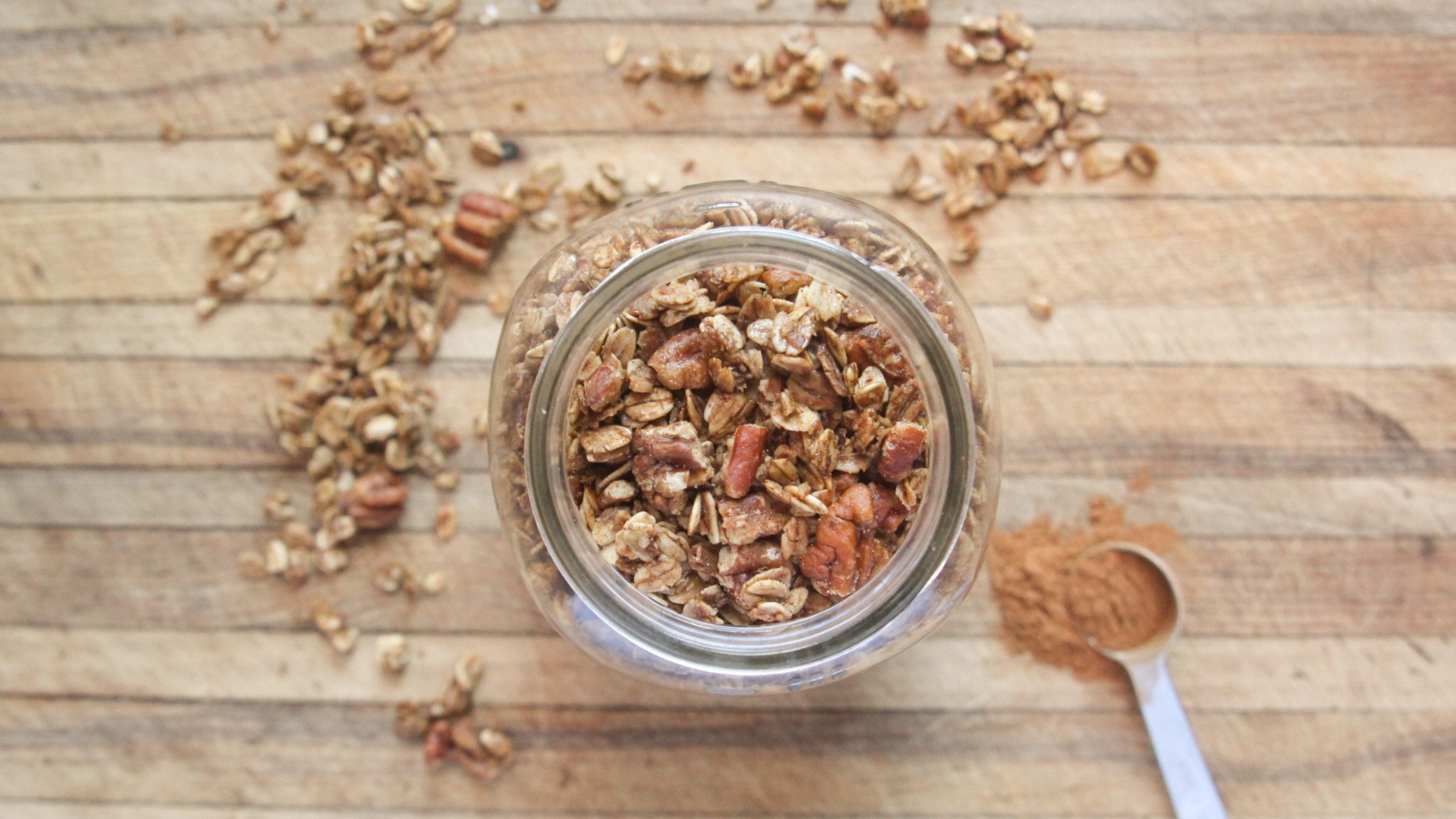 Top-down view of homemade pecan pie granola on a cutting board with a teaspoon of cinnamon and scattered granola.