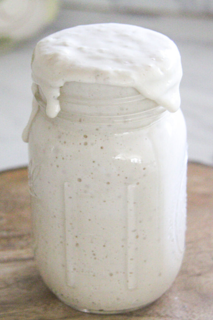 A glass mason jar overflowing with active, bubbly white sourdough starter on a wooden surface.