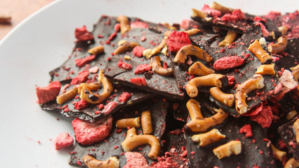 Strawberry Pretzel Bark (Dairy-Free) A close-up, angled shot of a pile of vegan strawberry pretzel chocolate bark on a white ceramic plate.