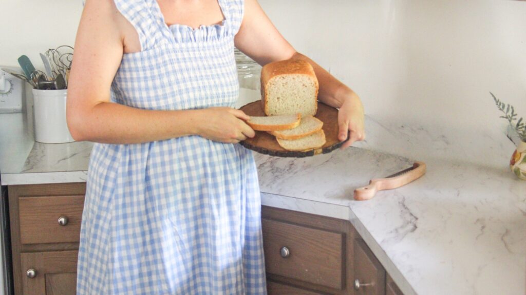 A woman in a blue gingham dress holding a wooden board with a sliced sourdough loaf in a bright, farmhouse-style kitchen.