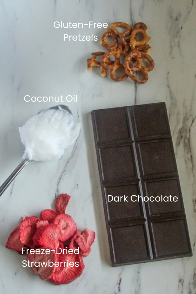 Strawberry Pretzel Bark (Dairy-Free) A marble surface displaying the four ingredients for the recipe: gluten-free pretzels, a bar of dark chocolate, coconut oil on a spoon, and freeze-dried strawberries.