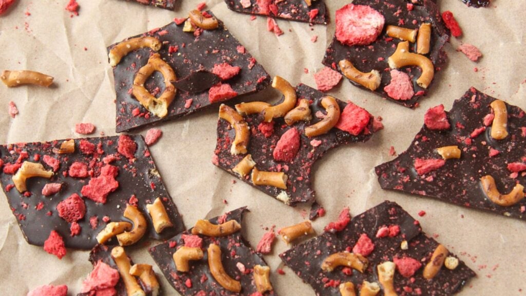 Flat lay of broken shards of dark chocolate bark topped with crushed pretzels and freeze-dried strawberries on brown parchment paper.