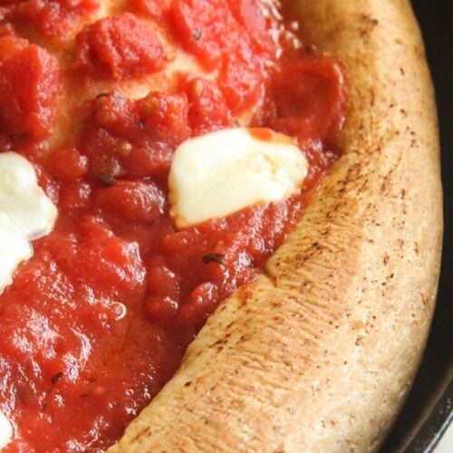 A close-up of a thick-crust spelt pizza with red tomato sauce and fresh mozzarella pearls baking in a cast iron skillet.