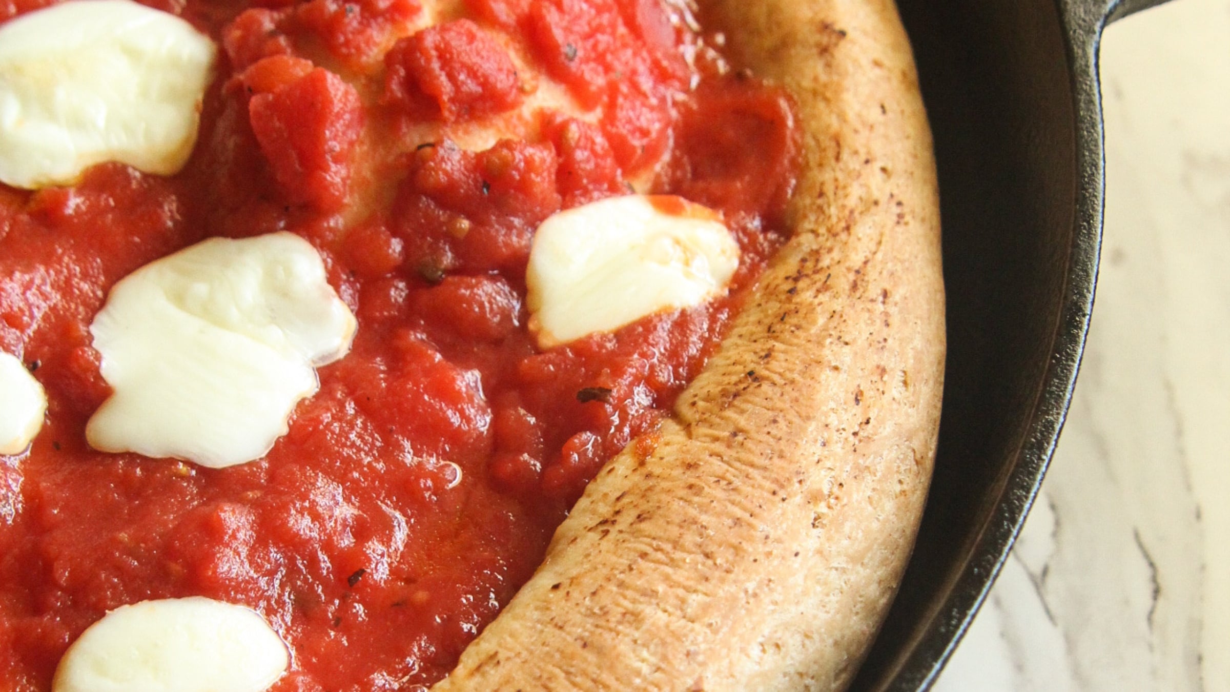 A close-up of a thick-crust spelt pizza with red tomato sauce and fresh mozzarella pearls baking in a cast iron skillet.