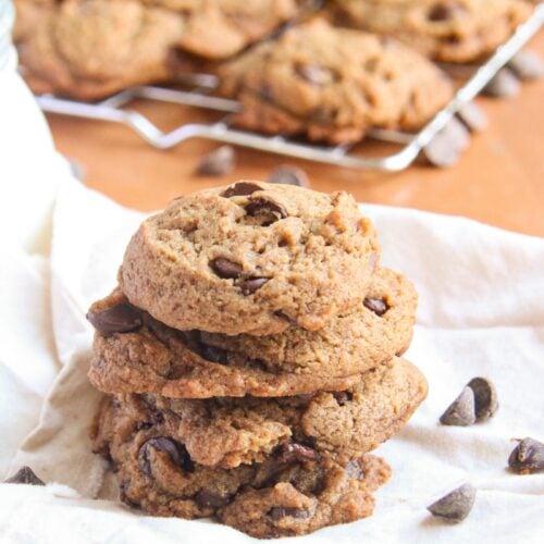 A stack of three thick chocolate chip cookies made with coconut sugar next to a glass of milk and scattered chocolate chips.