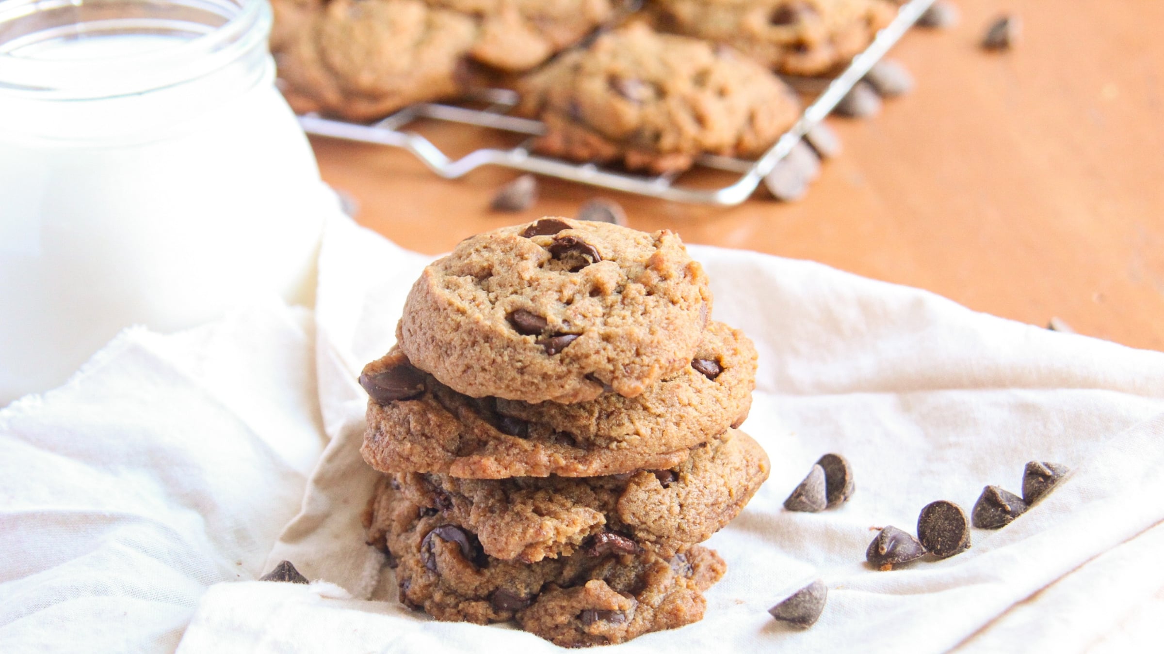 A stack of three thick chocolate chip cookies made with coconut sugar next to a glass of milk and scattered chocolate chips.