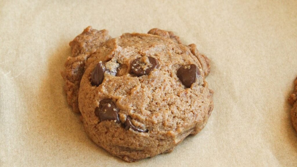 A single baked chocolate chip cookie sitting on brown parchment paper with visible sea salt flakes on top.