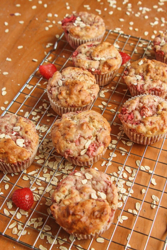 Vertical view of strawberry oatmeal muffins cooling on a rack with scattered oats and fresh strawberries on a wooden table.