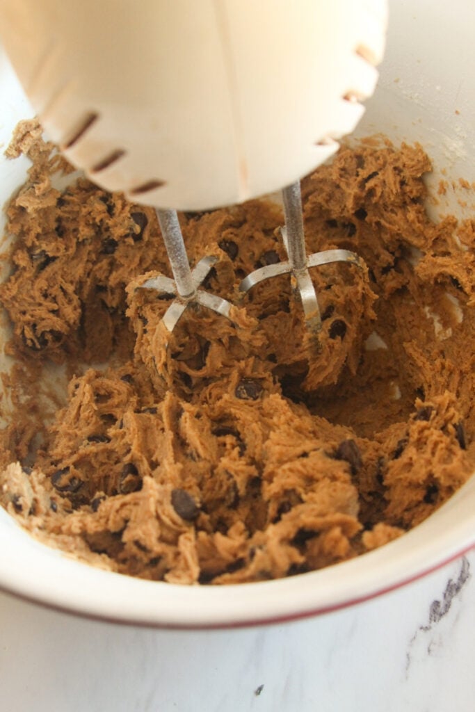 An electric hand mixer blending thick cookie dough with dark chocolate chips in a white mixing bowl.