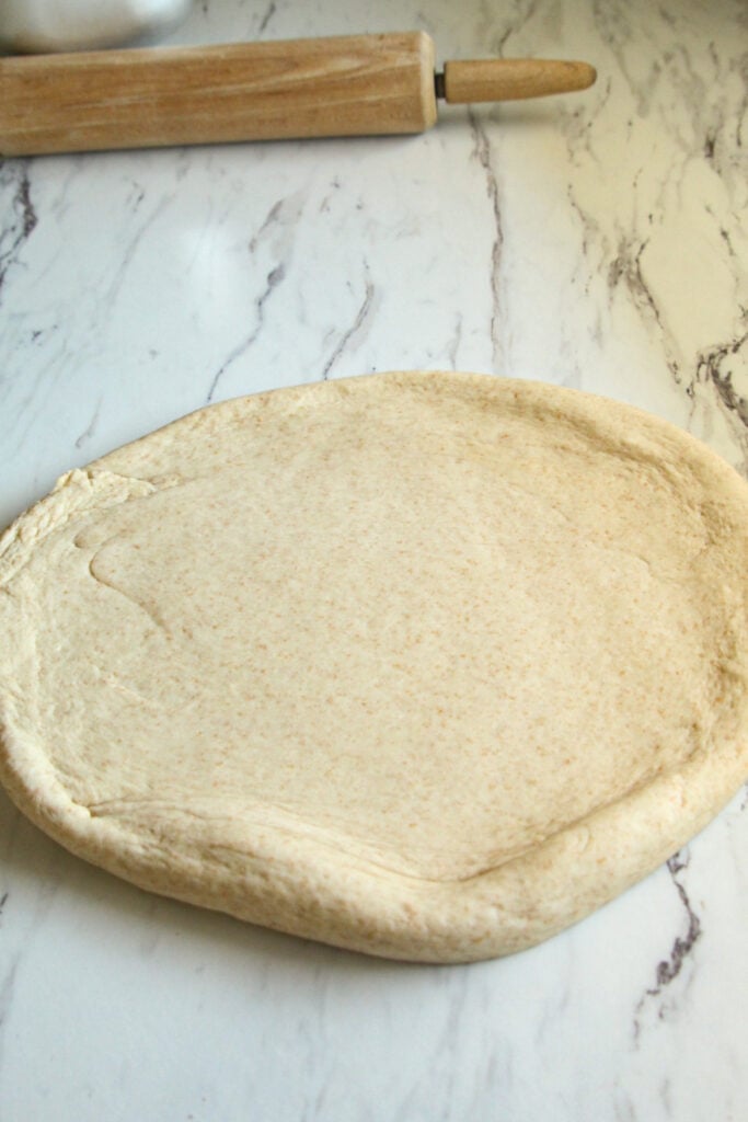 Unbaked spelt pizza dough rolled into a circle on a white marble countertop next to a wooden rolling pin.