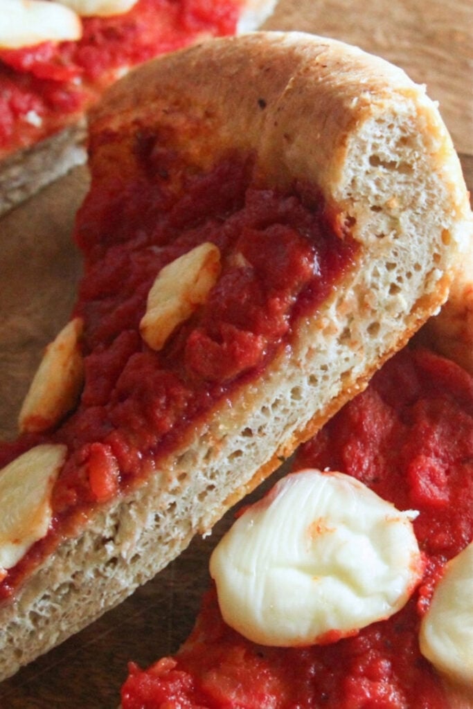 A vertical close-up of a slice of spelt pizza showing the airy crumb of the crust and melted cheese on top.